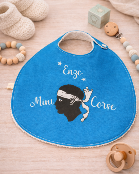 Bavoir bébé personnalisé Mini Corse avec prénom Enzo posé sur une table avec accessoires bébé, création artisanale