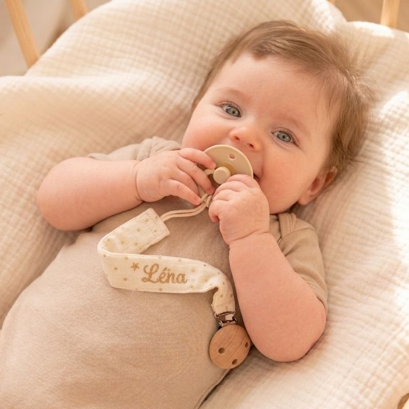 Bébé portant une attache tétine personnalisée en tissu double gaze avec prénom, fixée au body pour éviter la perte de la tétine.